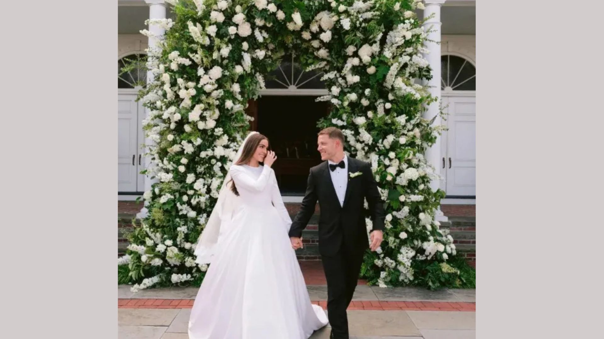 "A bride in a flowing white gown and a groom in a black tuxedo joyfully hold hands, walking past a lush floral arch of white blossoms."