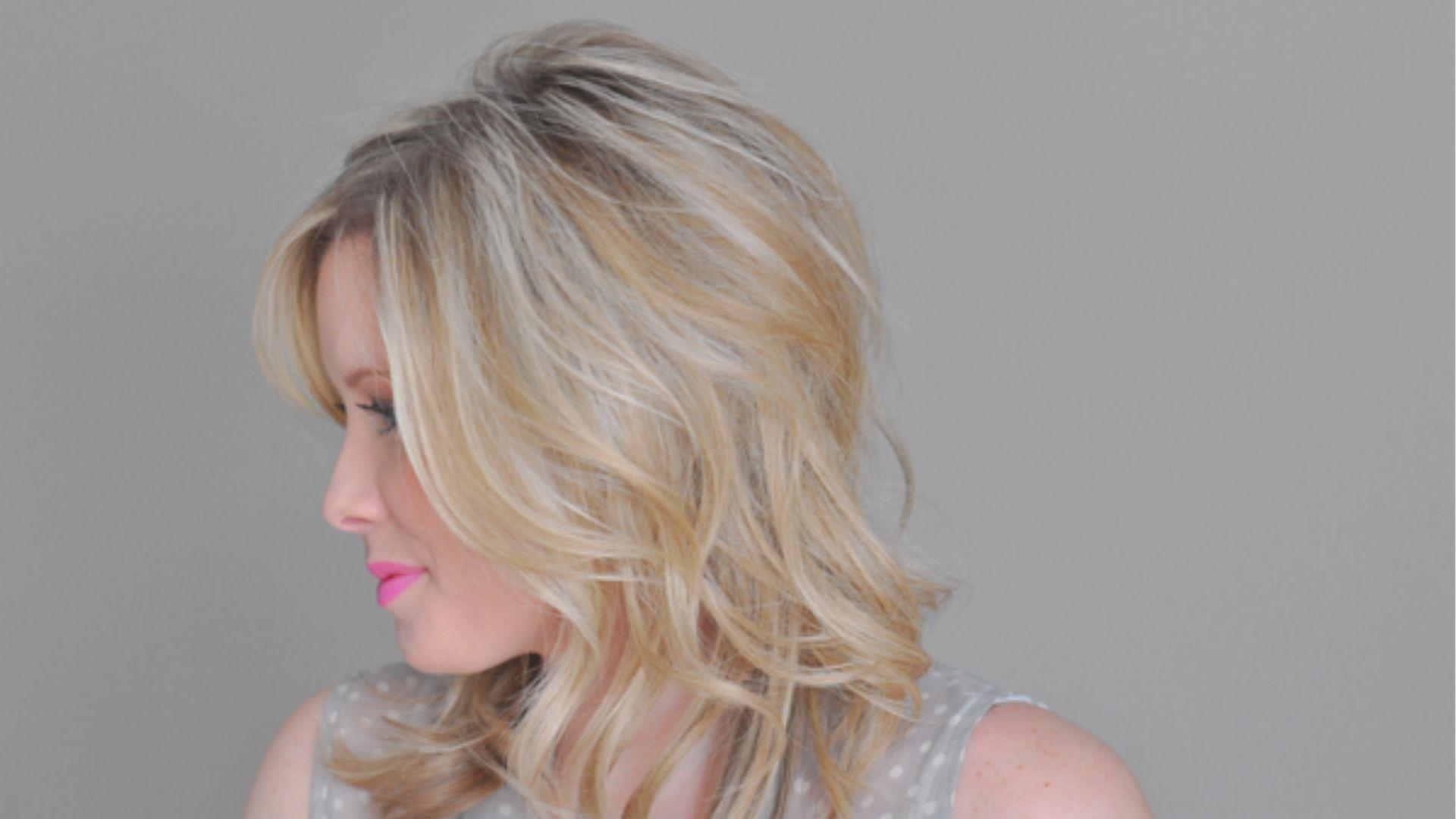 Side profile of a woman with soft, brushed-out blonde waves and curtain bangs.