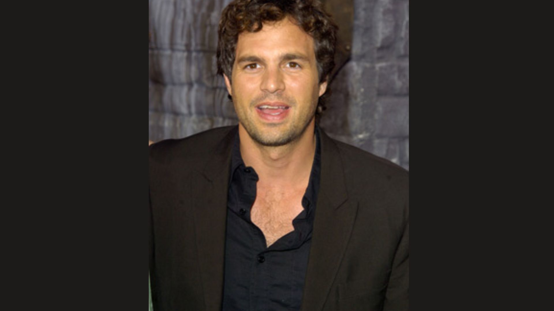 "A man with curly hair wearing a black shirt, standing and looking towards the camera."