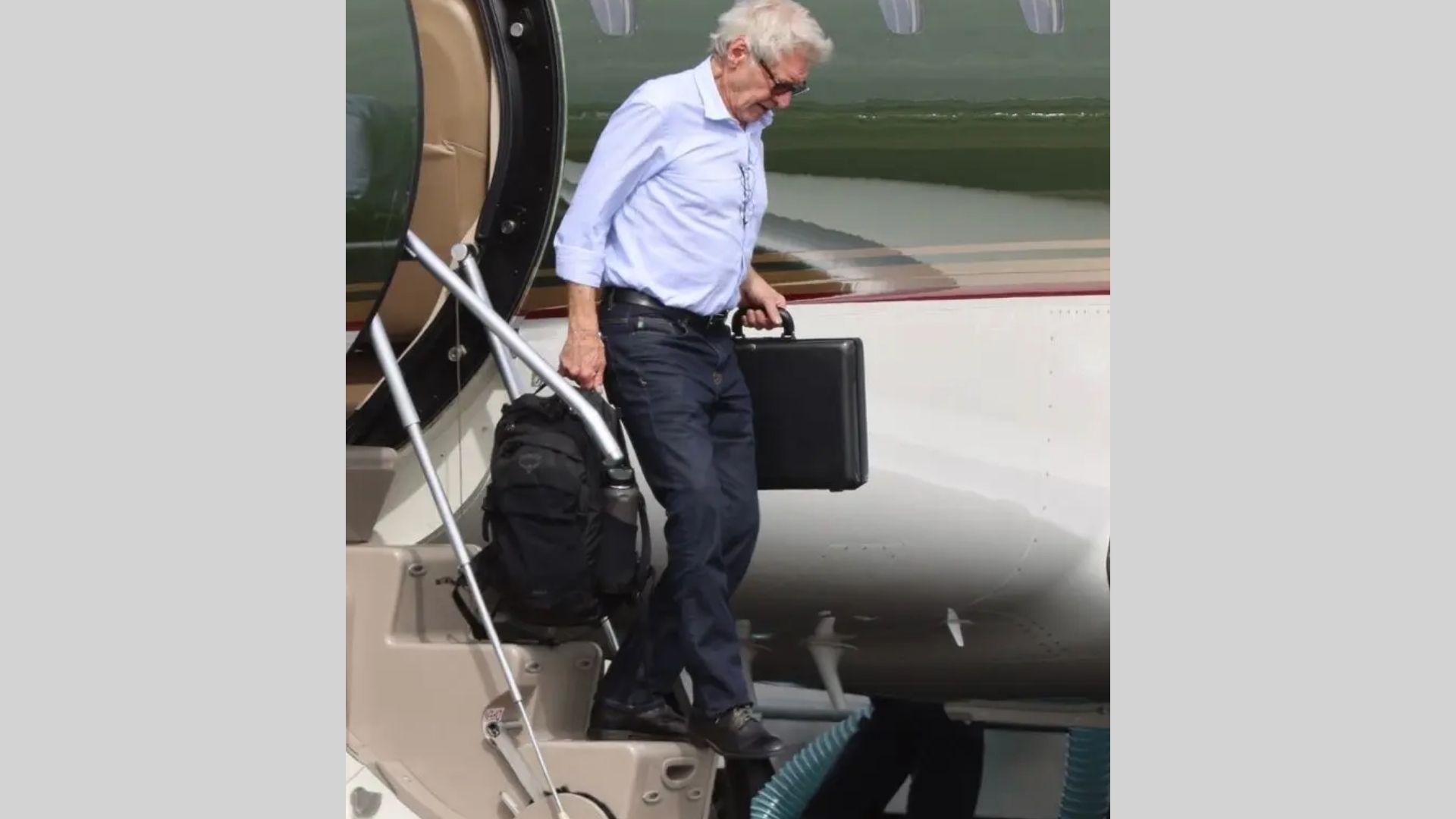 Older Harrison Ford exits private jet carrying briefcase and backpack in light blue shirt