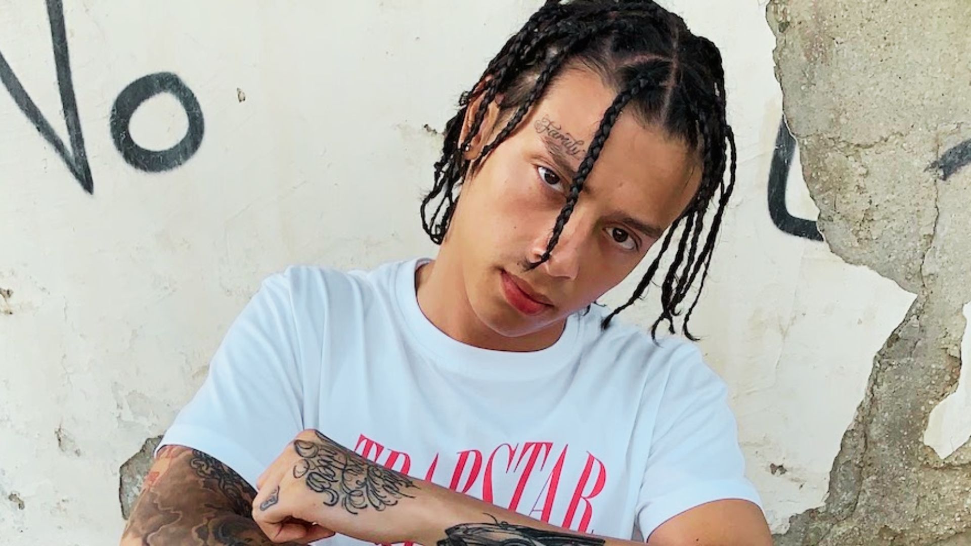 Central Cee poses against a cracked wall in a white tee, with shoulder-length box braids parted down the middle.