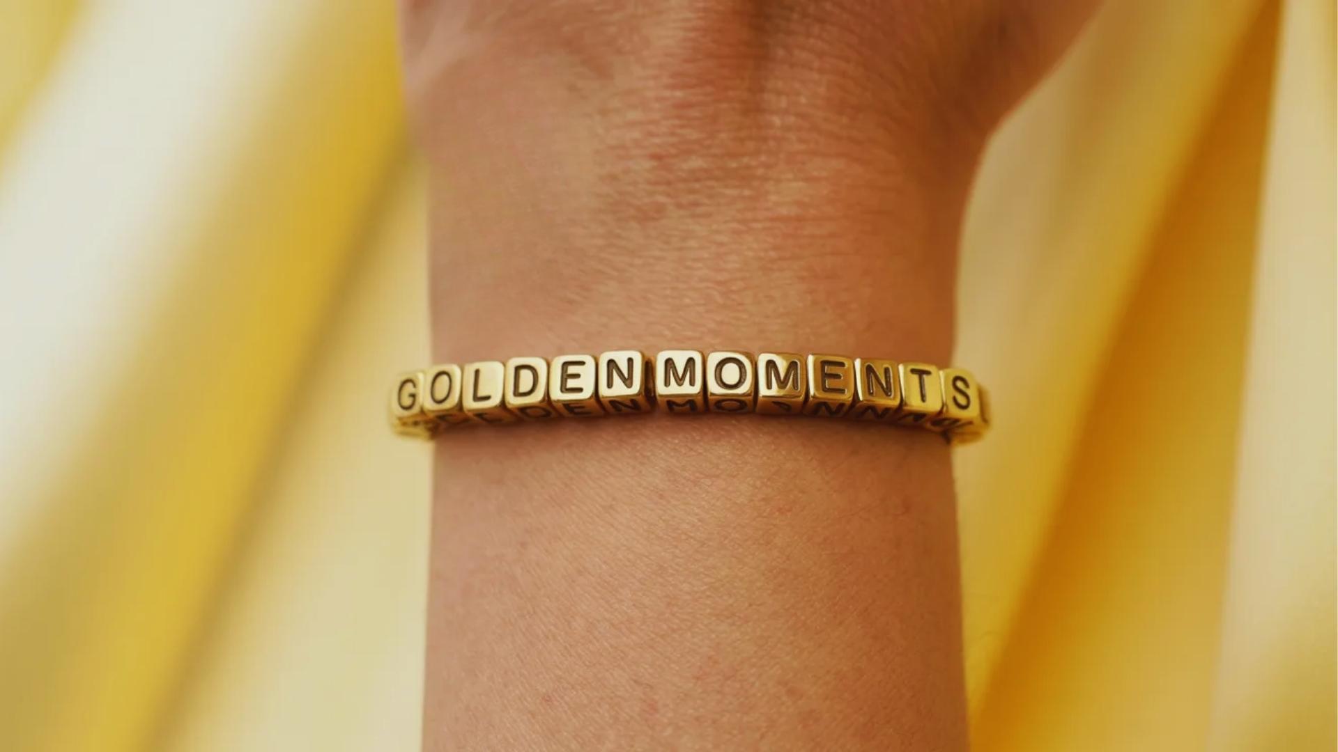 close-up wrist wearing a yellow and gold bracelet with cube beads spelling GOLDEN MOMENTS clearly in bold letters.