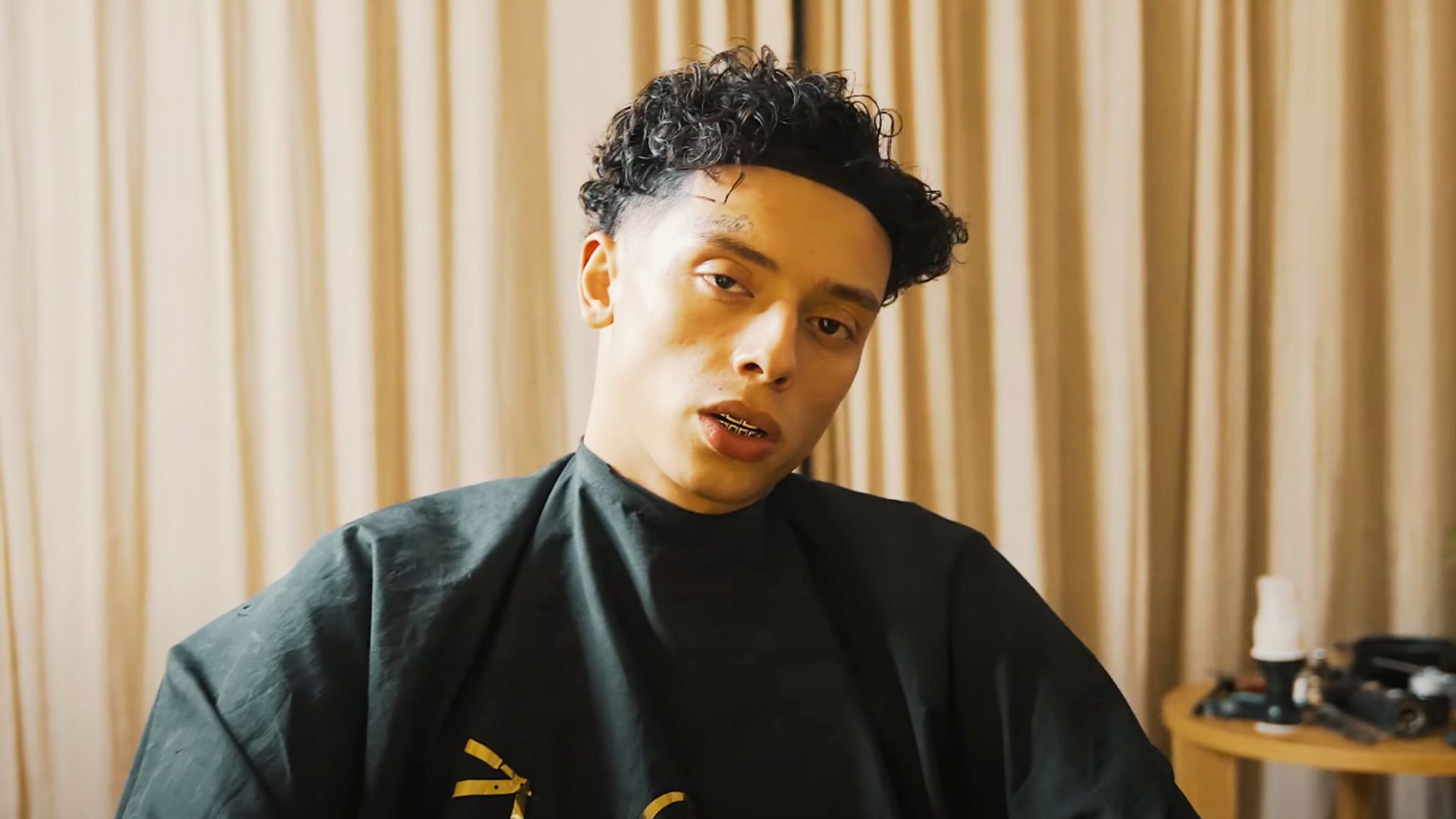 Central Cee sits in a barber cape mid-cut, showing off his signature curly top with a sharp skin fade.