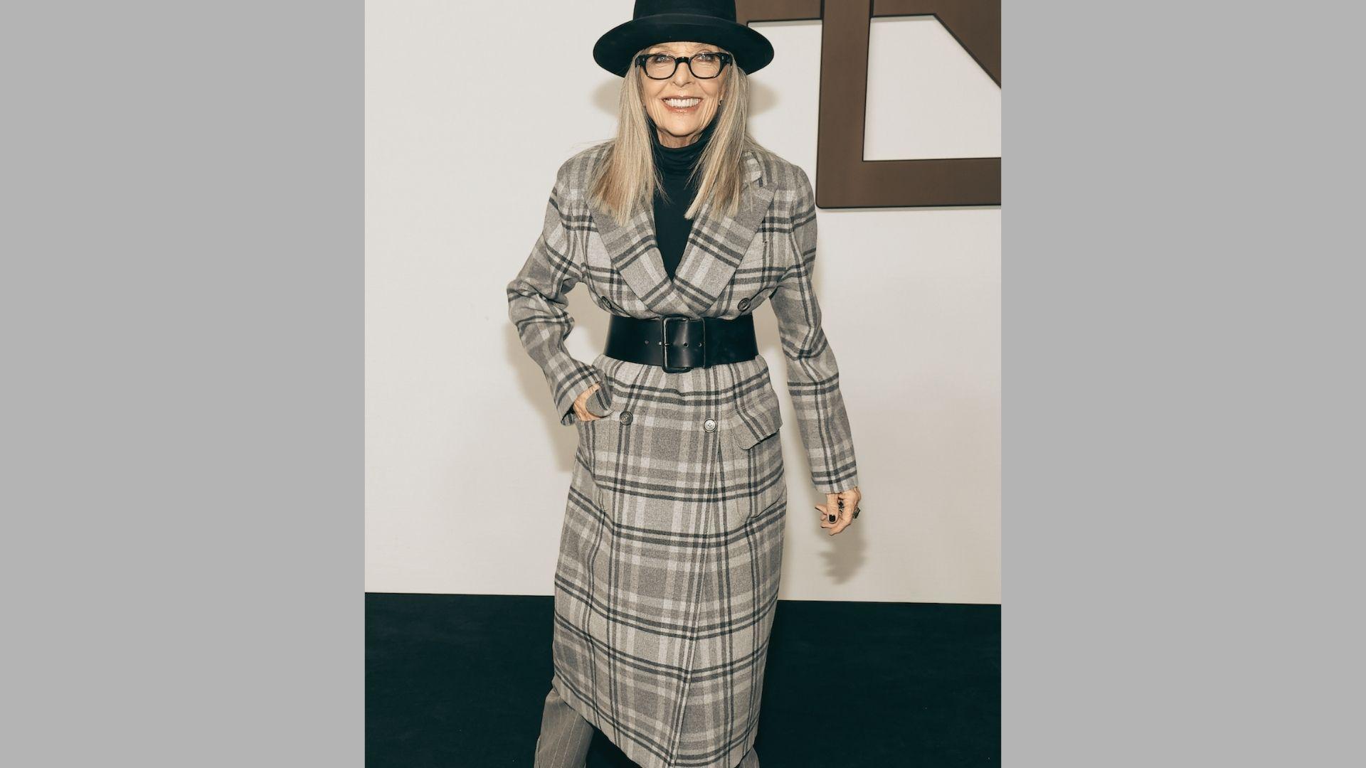 Woman in plaid belted coat and black hat poses confidently at fashion event backdrop