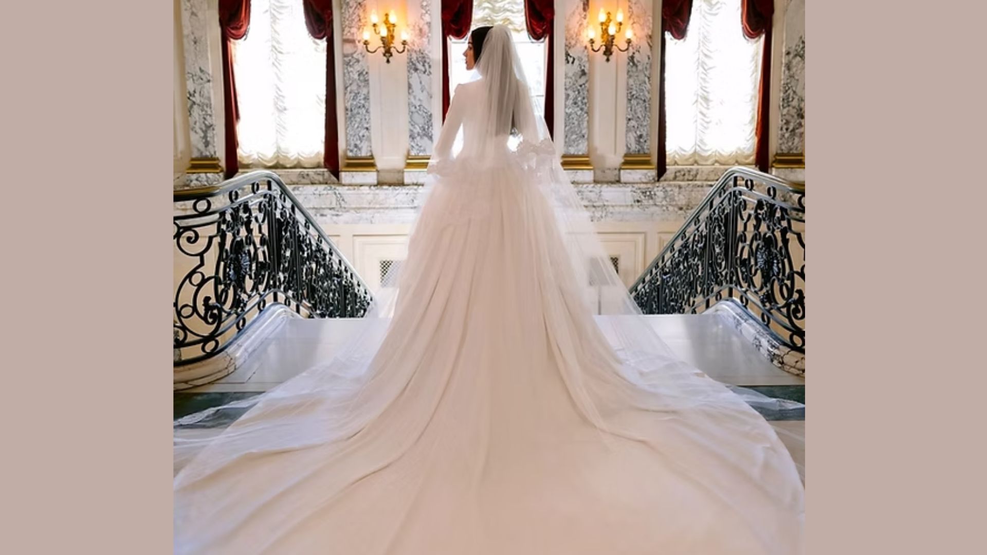 "A bride in a flowing white gown and veil stands atop a grand staircase adorned with ornate railings, under soft lighting from wall sconces."