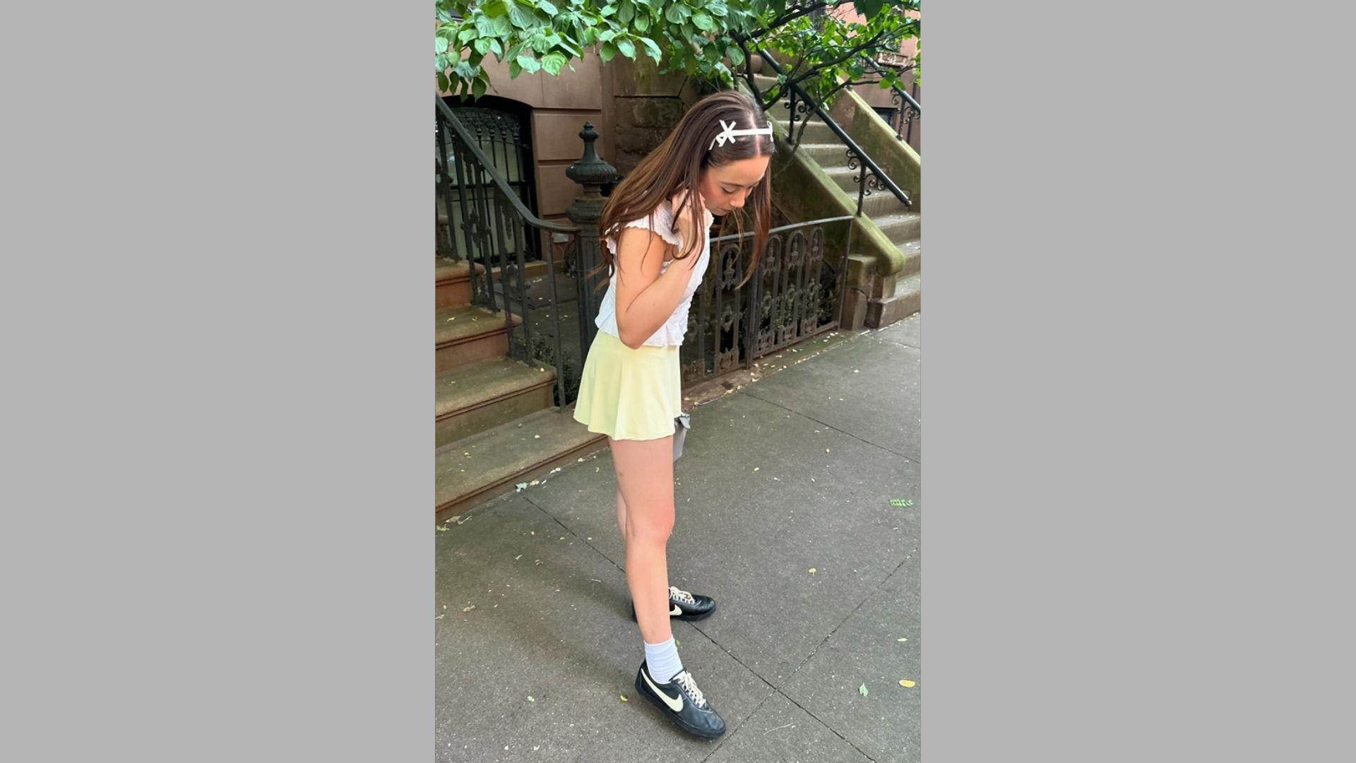 Young woman in white top and yellow skirt adjusts hair wearing sneakers on sidewalk