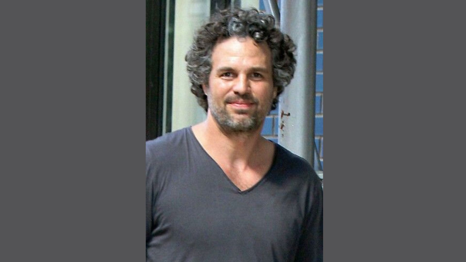 "A man with curly hair wearing a black shirt stands confidently, showcasing his casual style."