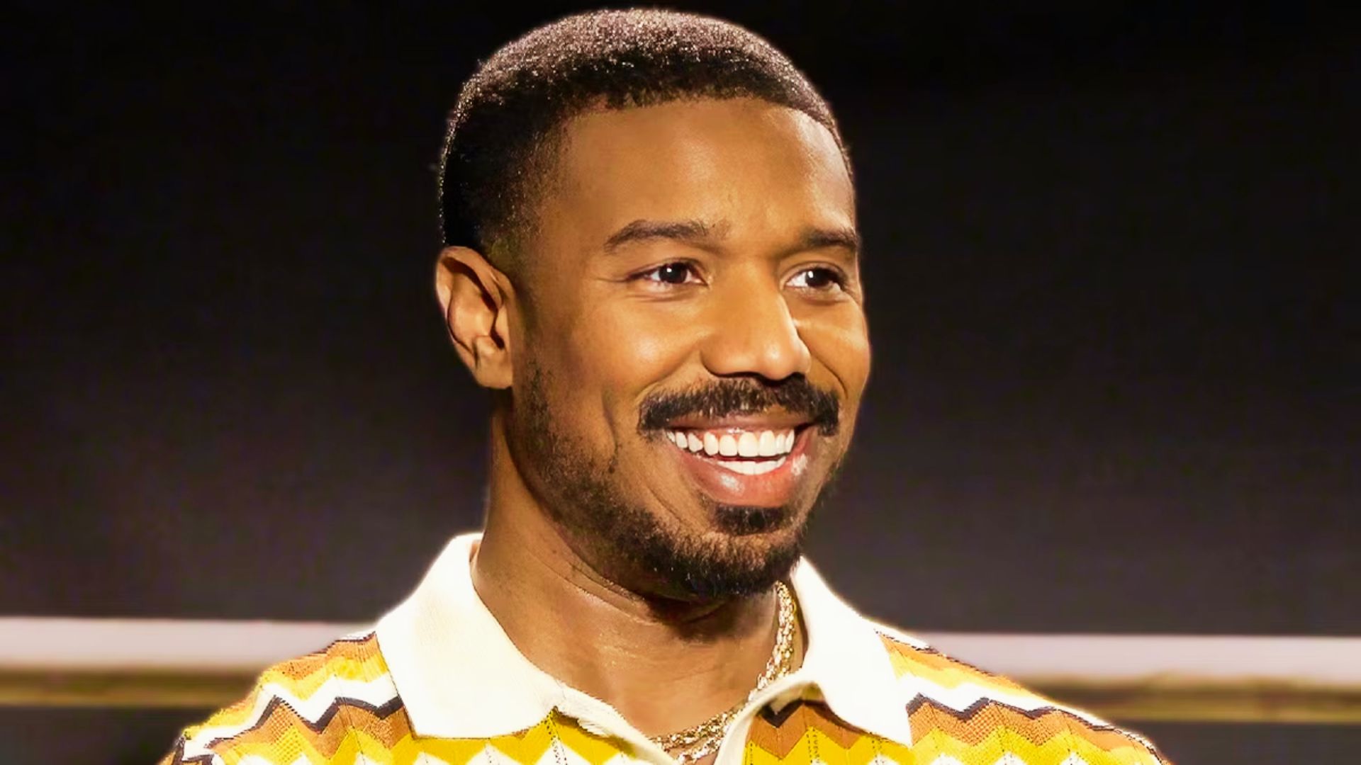 Michael B. Jordan smiling in a patterned polo, wearing a short buzz cut with a clean taper.