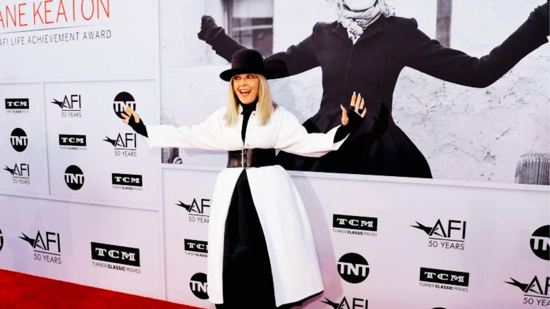 Woman in black bowler hat and white coat poses playfully on red carpet backdrop