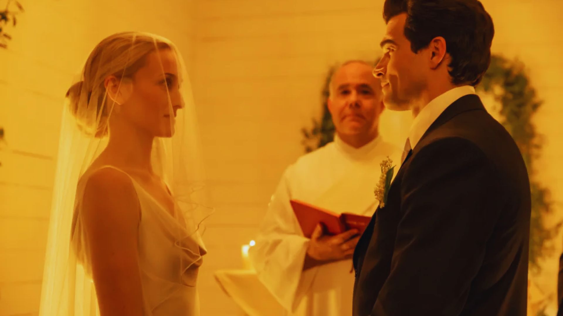 "A bride and groom face each other, standing in a warmly lit chapel. The bride wears a veil, while the groom is in a suit. A priest stands behind, holding a book."