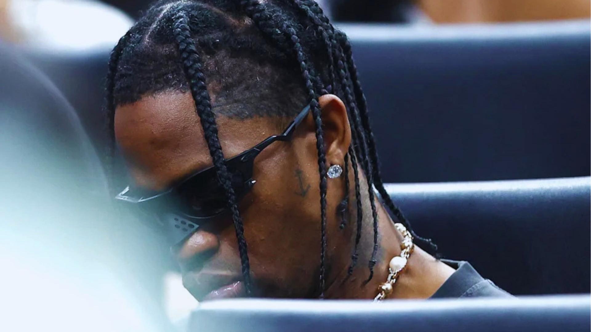 Close-up of Travis Scott with detailed cornrow parts and long box braids, wearing sunglasses and diamond earrings courtside.