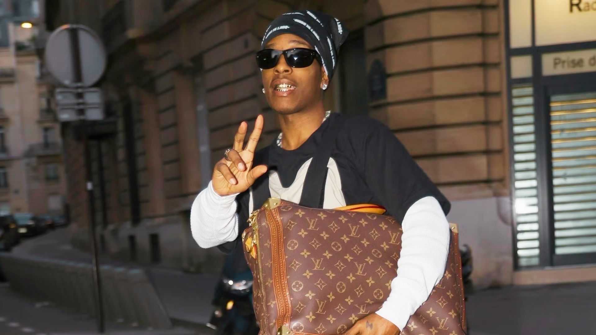 A$AP Rocky flashes a peace sign on a Paris street in layered streetwear, black bandana, and sunglasses.