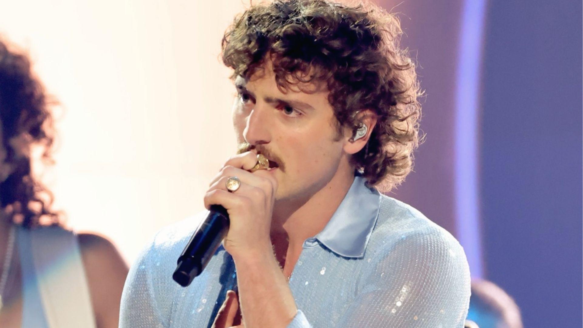 Benson Boone sings in a light blue shirt showing fluffy voluminous curls and a mustache.