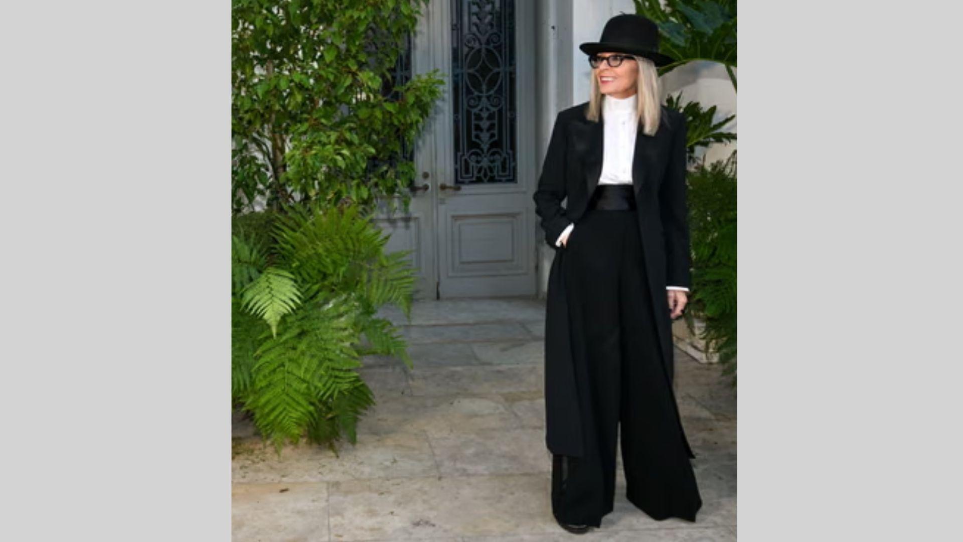 Woman in black coat, wide trousers, and hat stands smiling outdoors by gray door