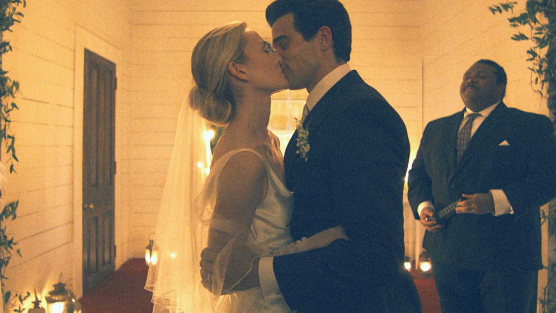 "A bride and groom share a kiss in a warmly lit corridor adorned with candles, while an officiant stands behind them, conveying a romantic atmosphere."