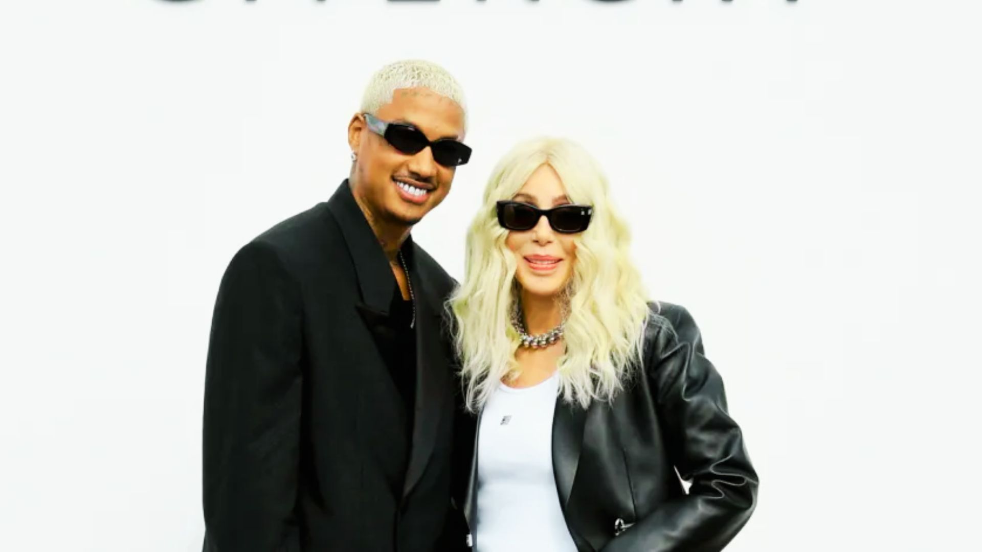 Cher with platinum blonde hair and dark sunglasses, posing in a black leather jacket over a white top.