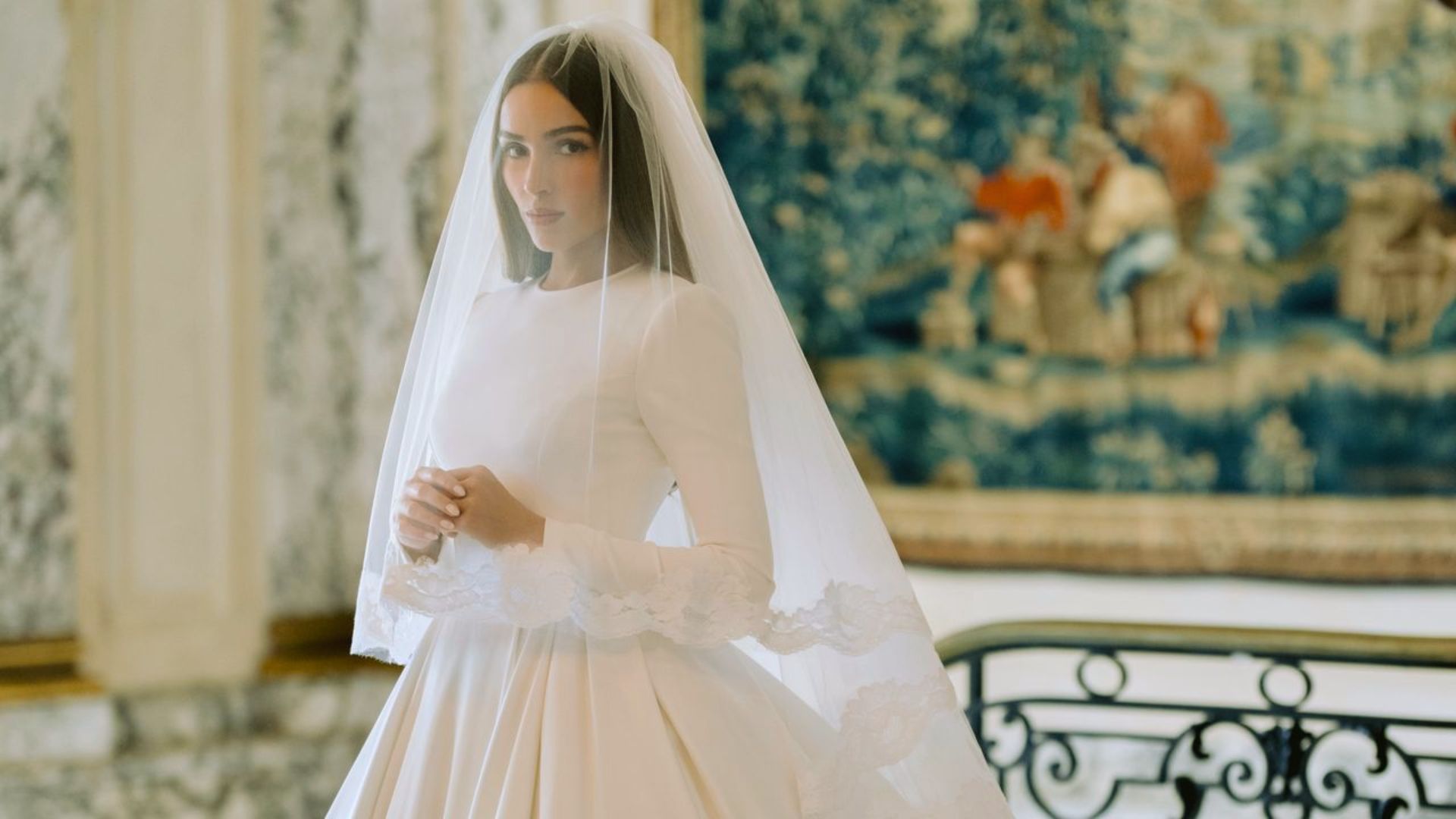"Bride in a classic white gown with a long veil stands gracefully. The backdrop features an ornate blue tapestry, creating a serene atmosphere."