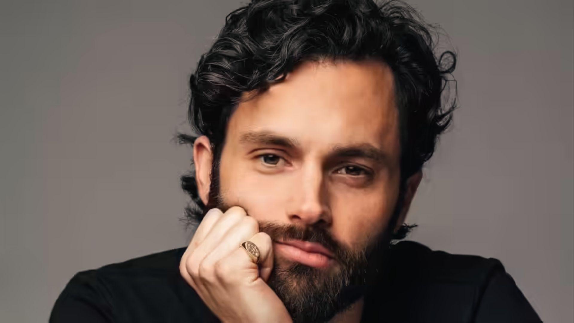 Penn Badgley poses with tousled dark curls and trimmed beard, hand on chin, gray background.
