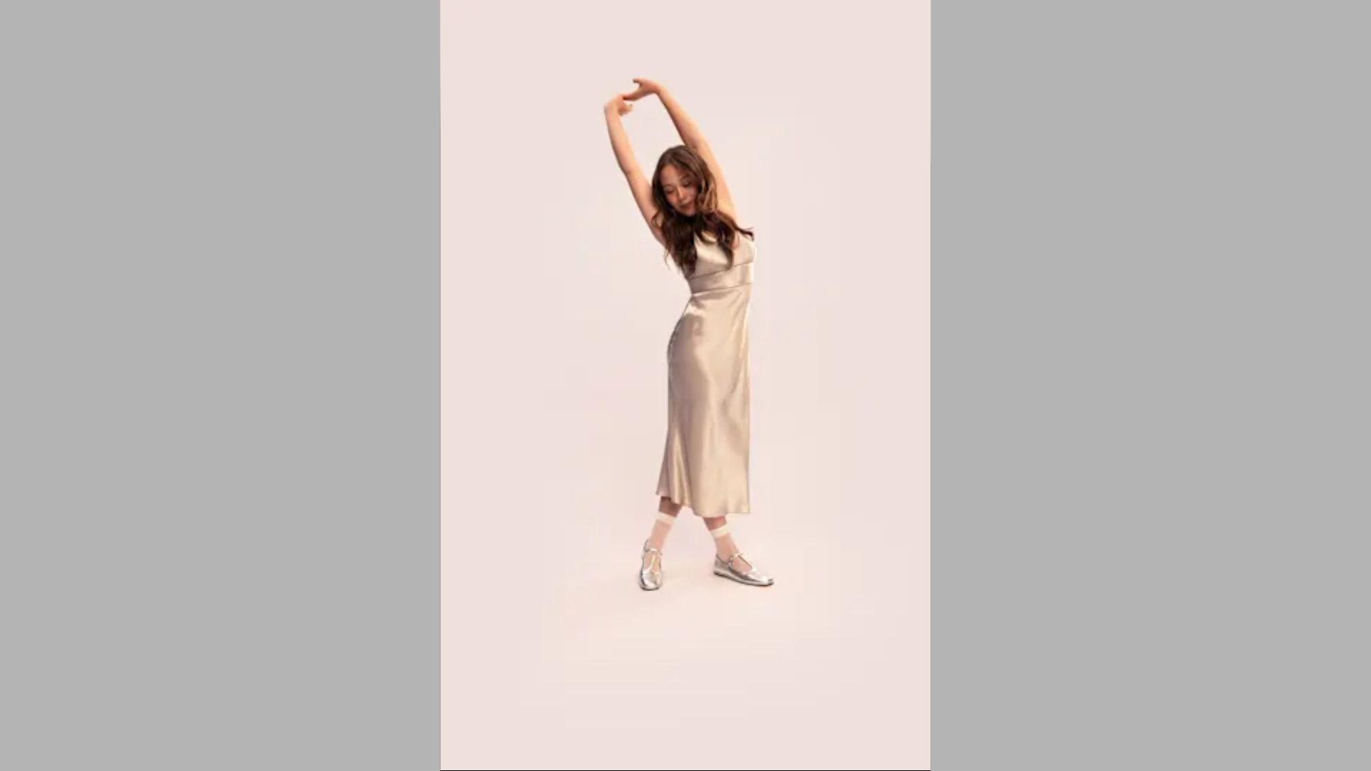 Woman in champagne satin dress stretches arms upward wearing silver flats against soft studio background