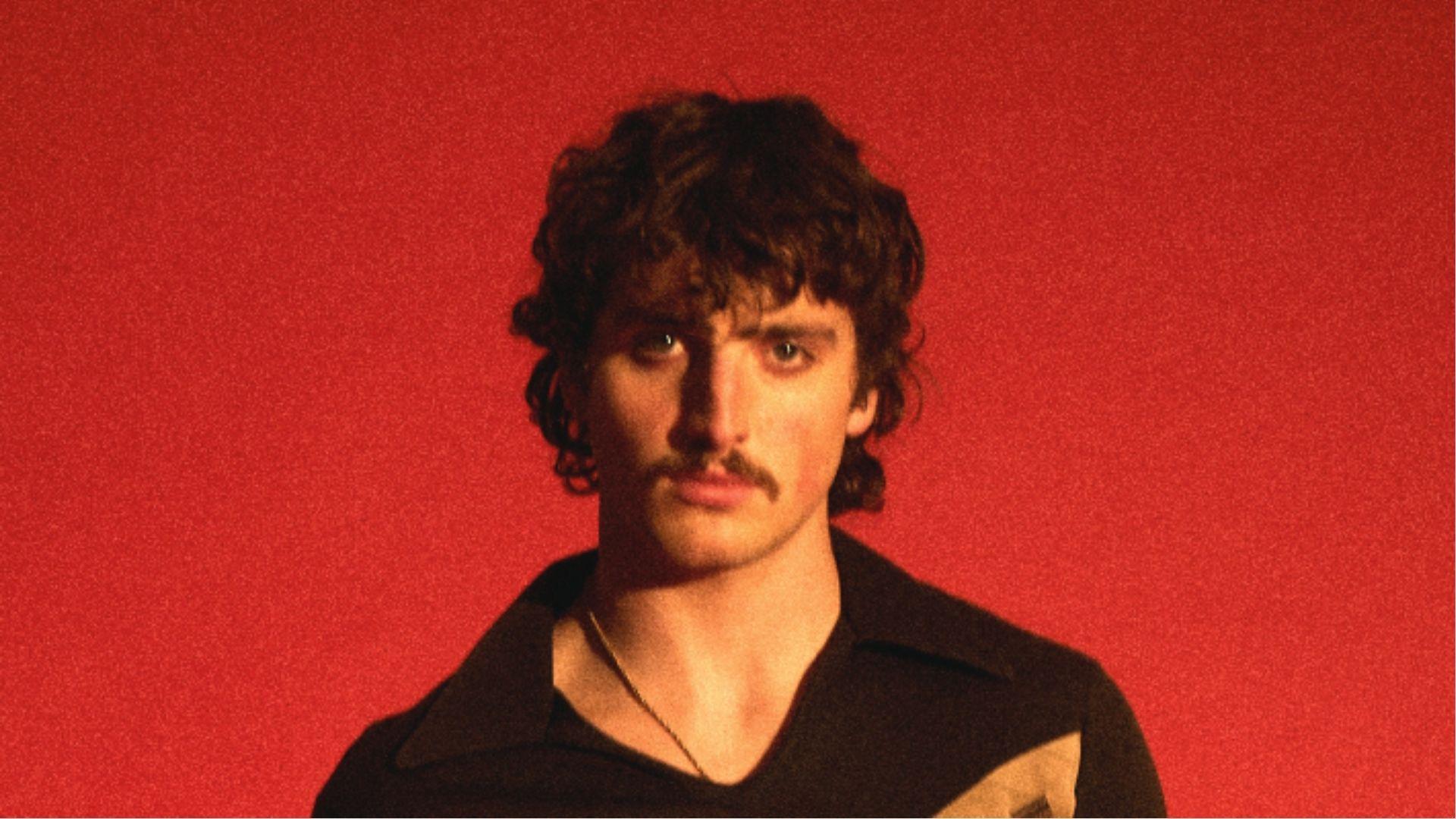 Benson Boone on a red backdrop sporting shaggy curly mullet and thin mustache.