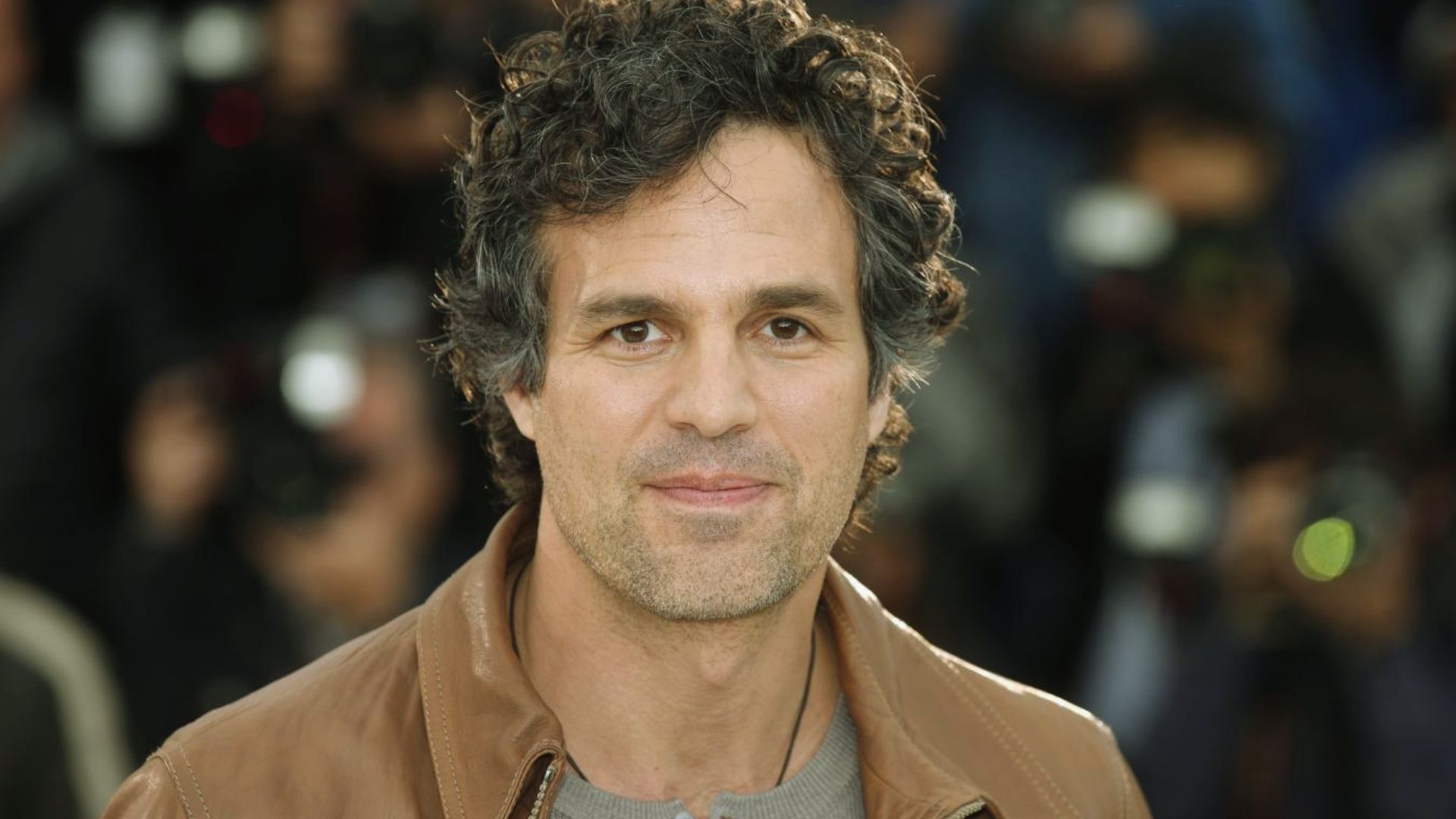 "A man with curly hair wearing a brown jacket stands, looking confidently at the camera."