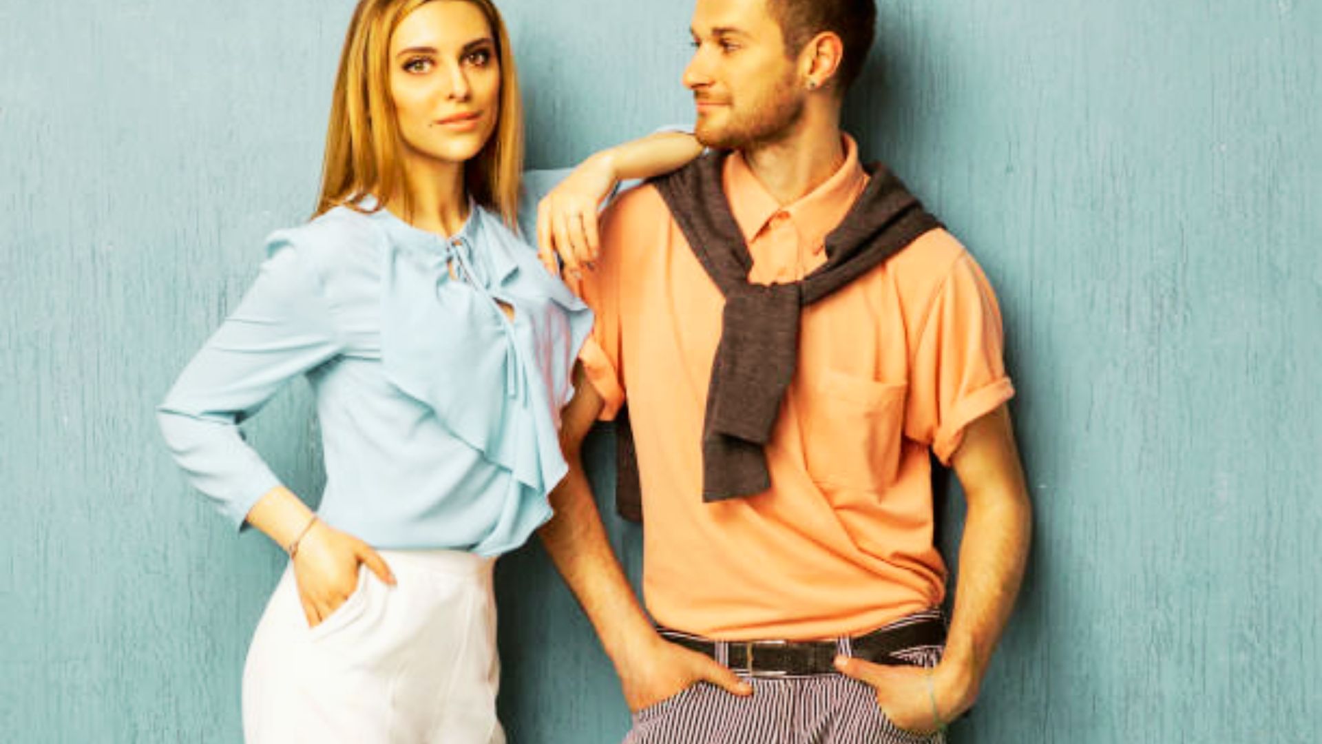 A stylish couple poses against a blue wall in complementary pastel outfits.