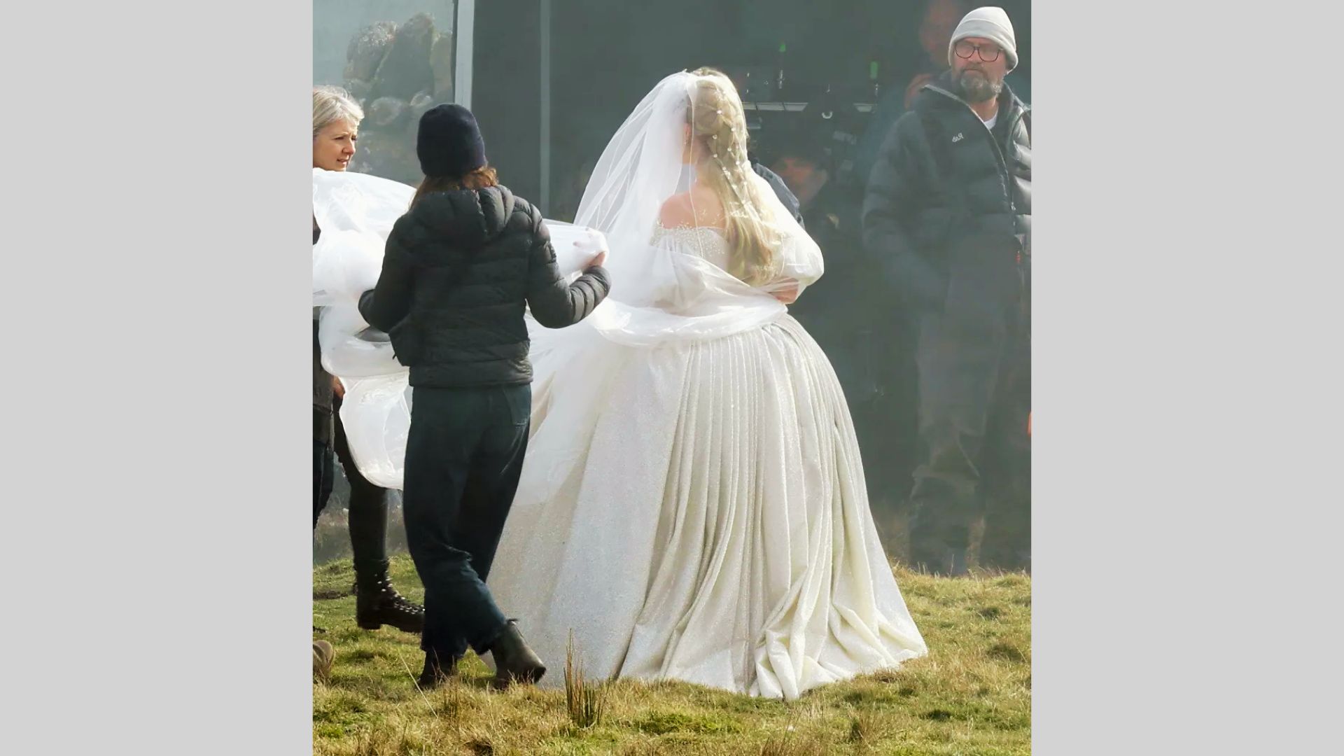 Crew members adjust veil of bride wearing white gown with pleated skirt on grassy set