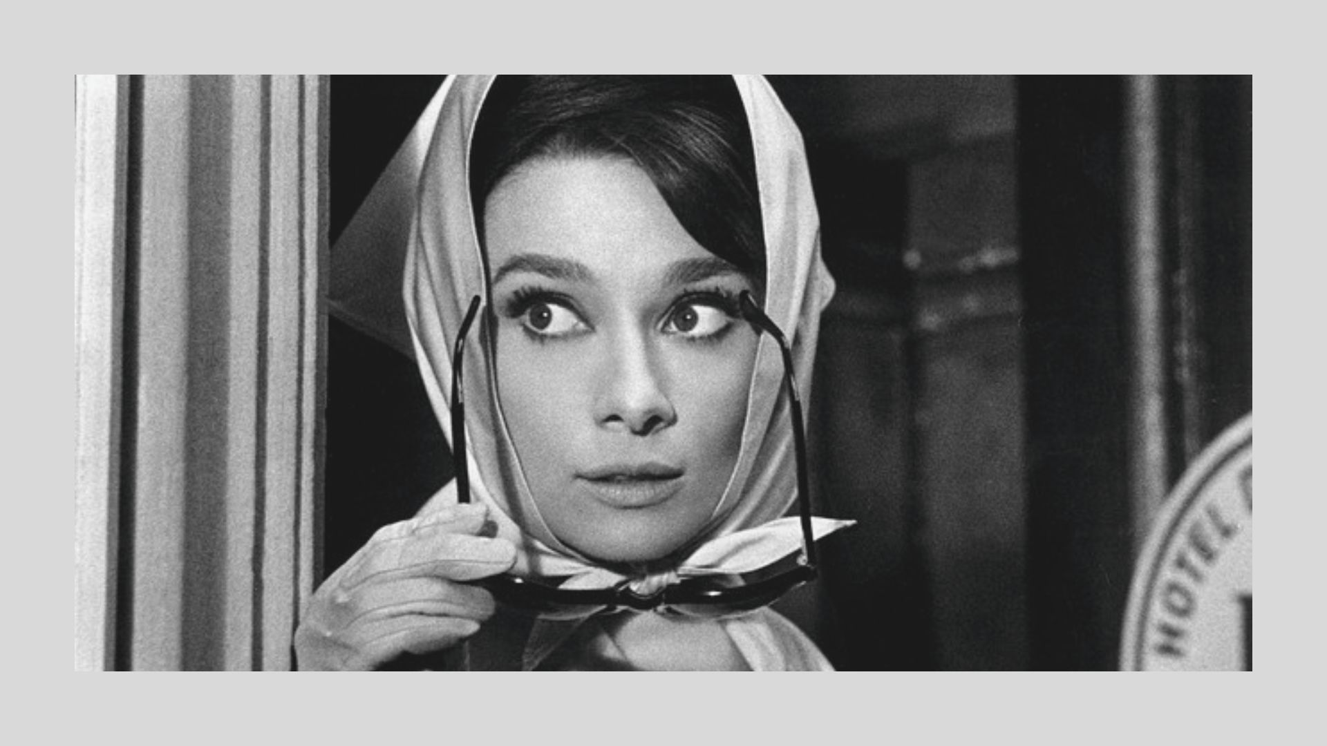 Black-and-white close-up of Audrey Hepburn in a white headscarf, white gloves, and holding cat-eye sunglasses.