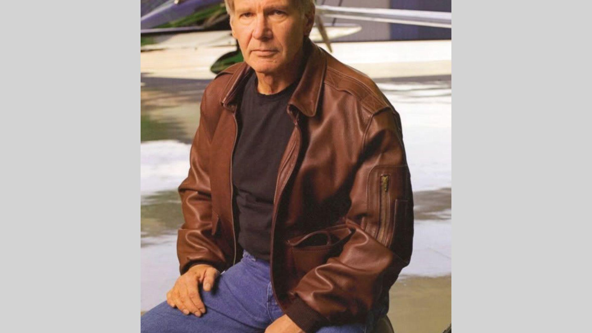 Harrison Ford sits wearing brown leather jacket and black shirt before private plane outdoors