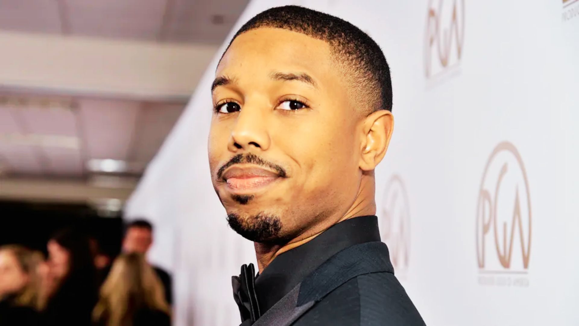 Michael B. Jordan on a red carpet in a black tuxedo, showing a clean buzz cut with a precise hairline.