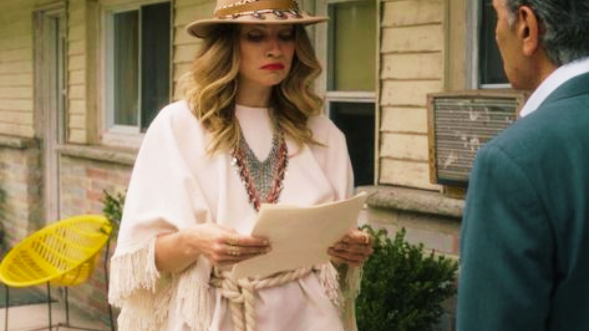 Alexis Rose stands outside the motel reading a paper in a cream fringed poncho cinched with a rope belt.