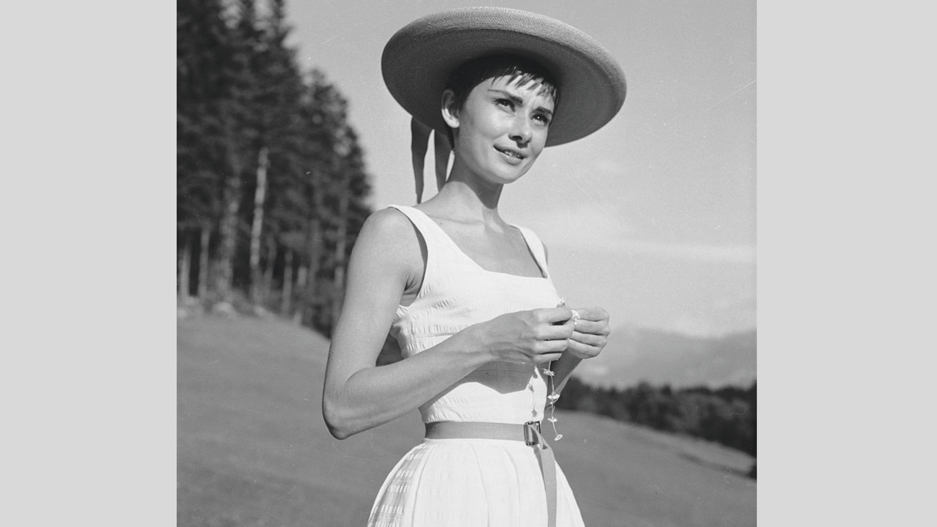 Audrey Hepburn outdoors in a wide-brim straw hat and square-neck sleeveless dress with a thin belt.