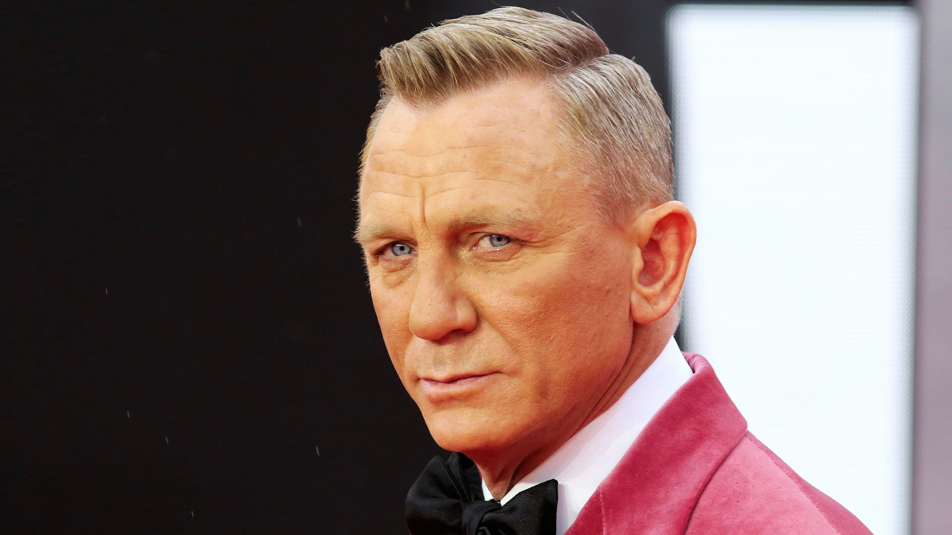 In a pink suit and bow tie, Daniel Craig presents a striking appearance, complementing his well-known James Bond hairstyle.