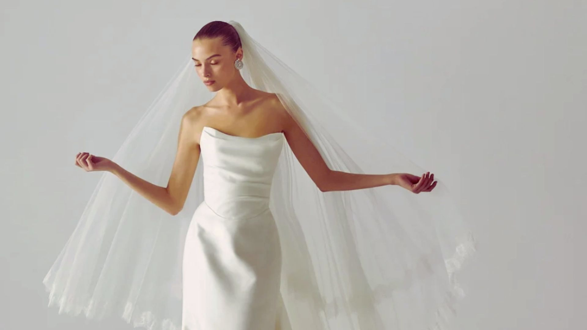 "A bride in a sleek, strapless wedding gown holds out her long, flowing veil with both hands, exuding style against a plain gray background."