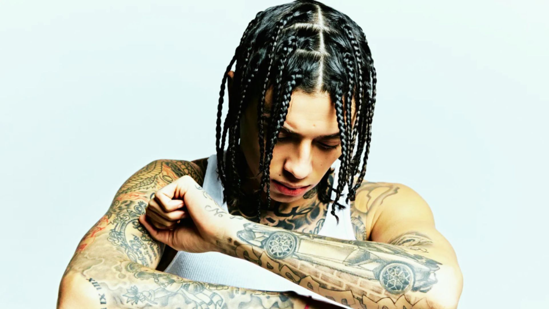 Central Cee looks down in a white tank top, featuring medium-length box braids with clean parts.