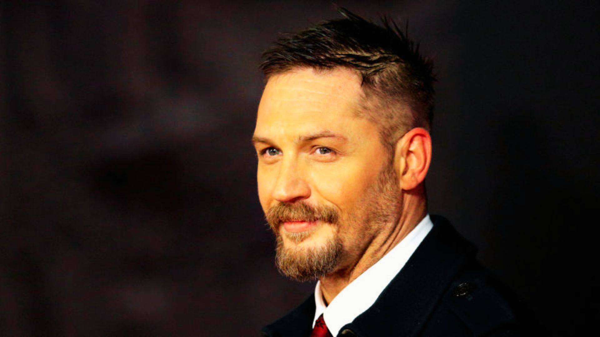 Tom Hardy with medium-length hair brushed back, faded sides, and light stubble.