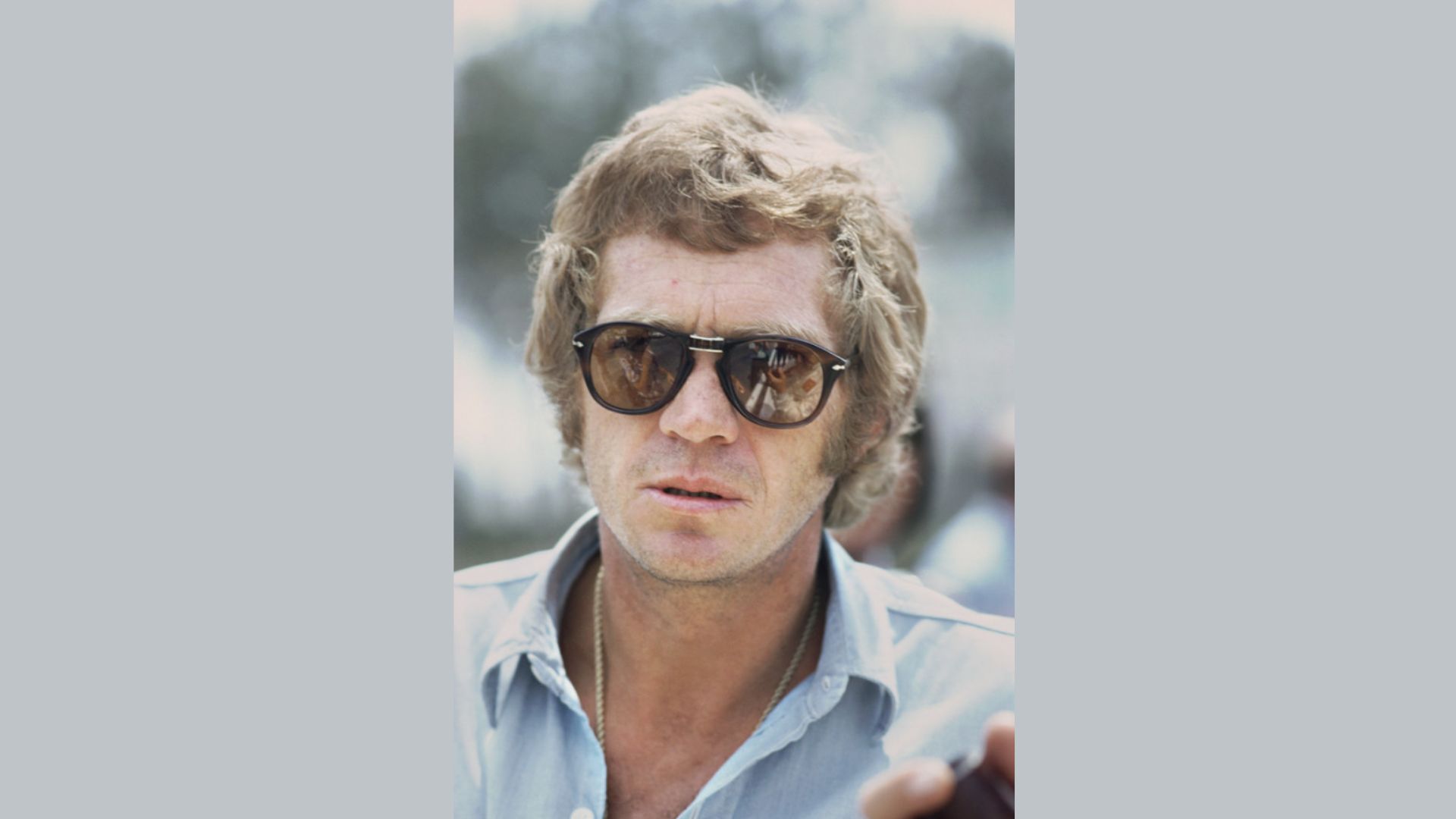 "A man wearing sunglasses and a blue shirt stands confidently, showcasing a casual and stylish look."