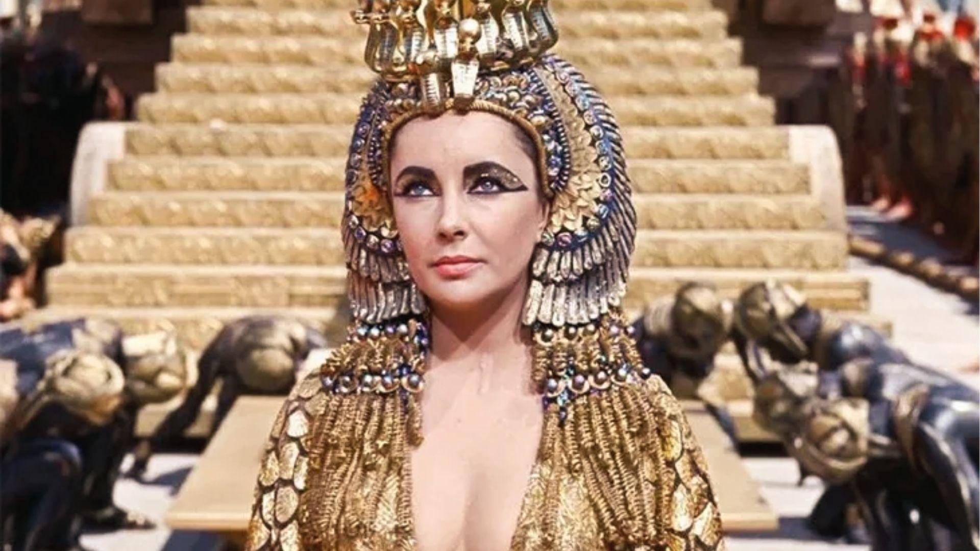 Elizabeth Taylor as Cleopatra in ornate gold headdress and gown before golden palace steps.