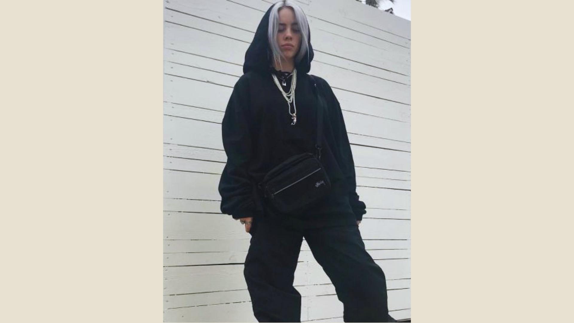 Billie Eilish in all-black oversized hoodie, baggy pants, crossbody bag, and layered necklaces with silver hair.