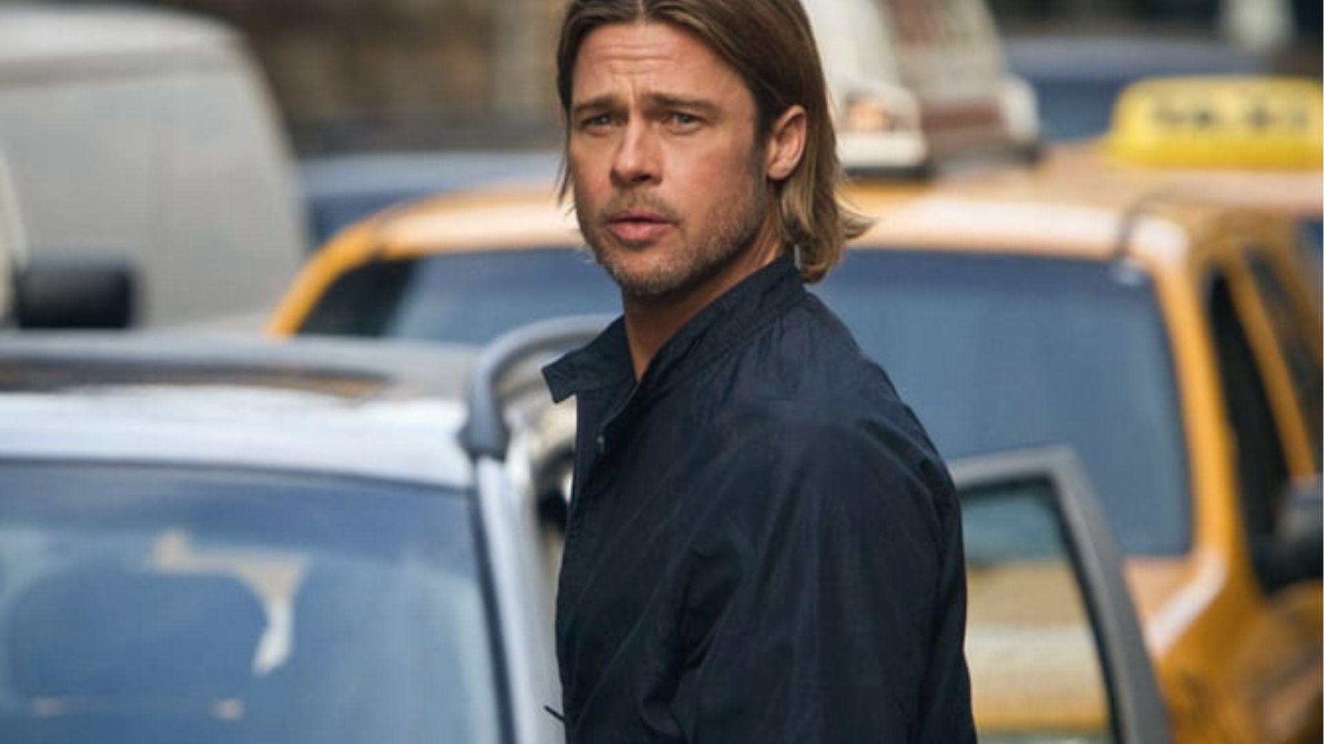 Brad Pitt with medium length hair on city street, dark jacket, yellow taxis behind