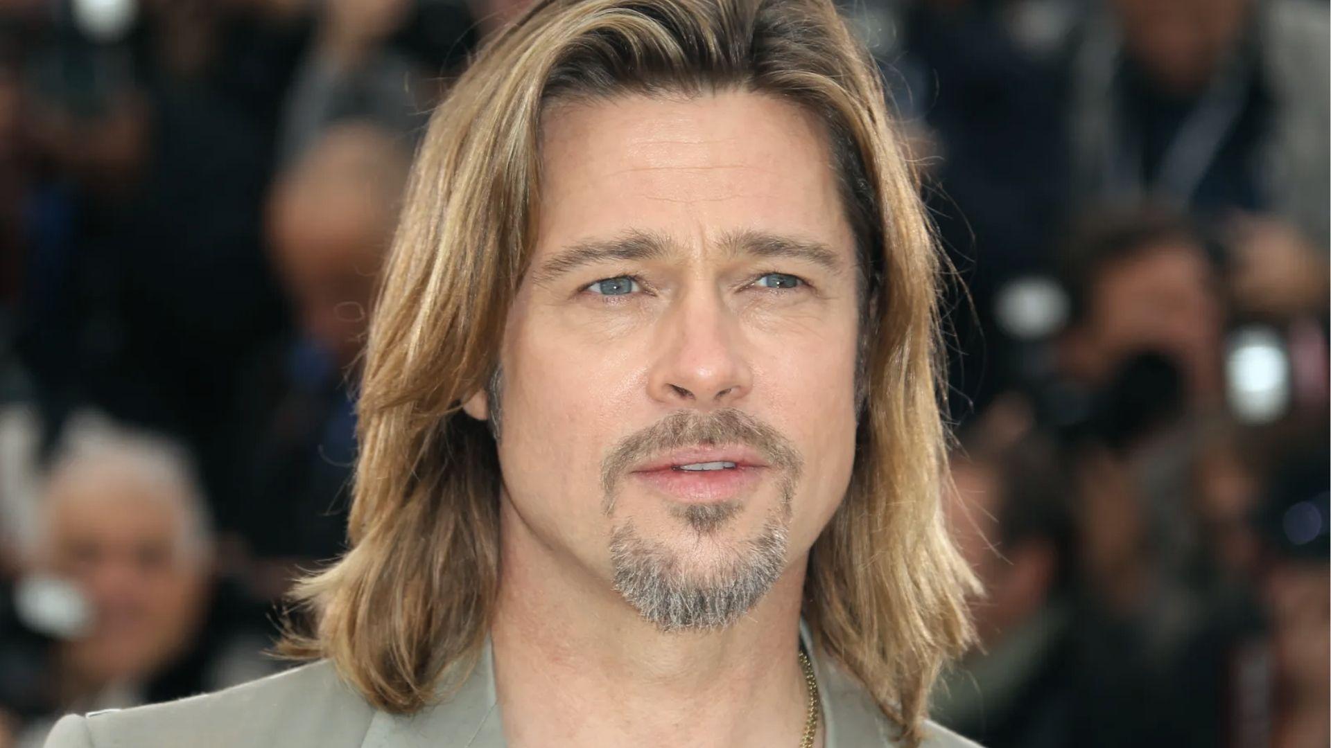 Brad Pitt with shoulder length blond hair and gray goatee at red carpet with photographers behind