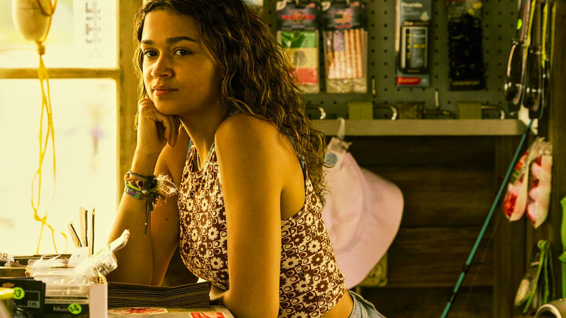 Kiara leans on a counter in a tackle shop wearing a brown floral print crop tank top and denim shorts.
