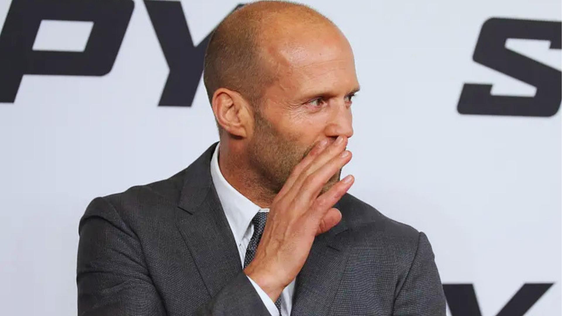 Jason Statham on the red carpet with a close buzz cut and salt-and-pepper beard, wearing a dark suit.