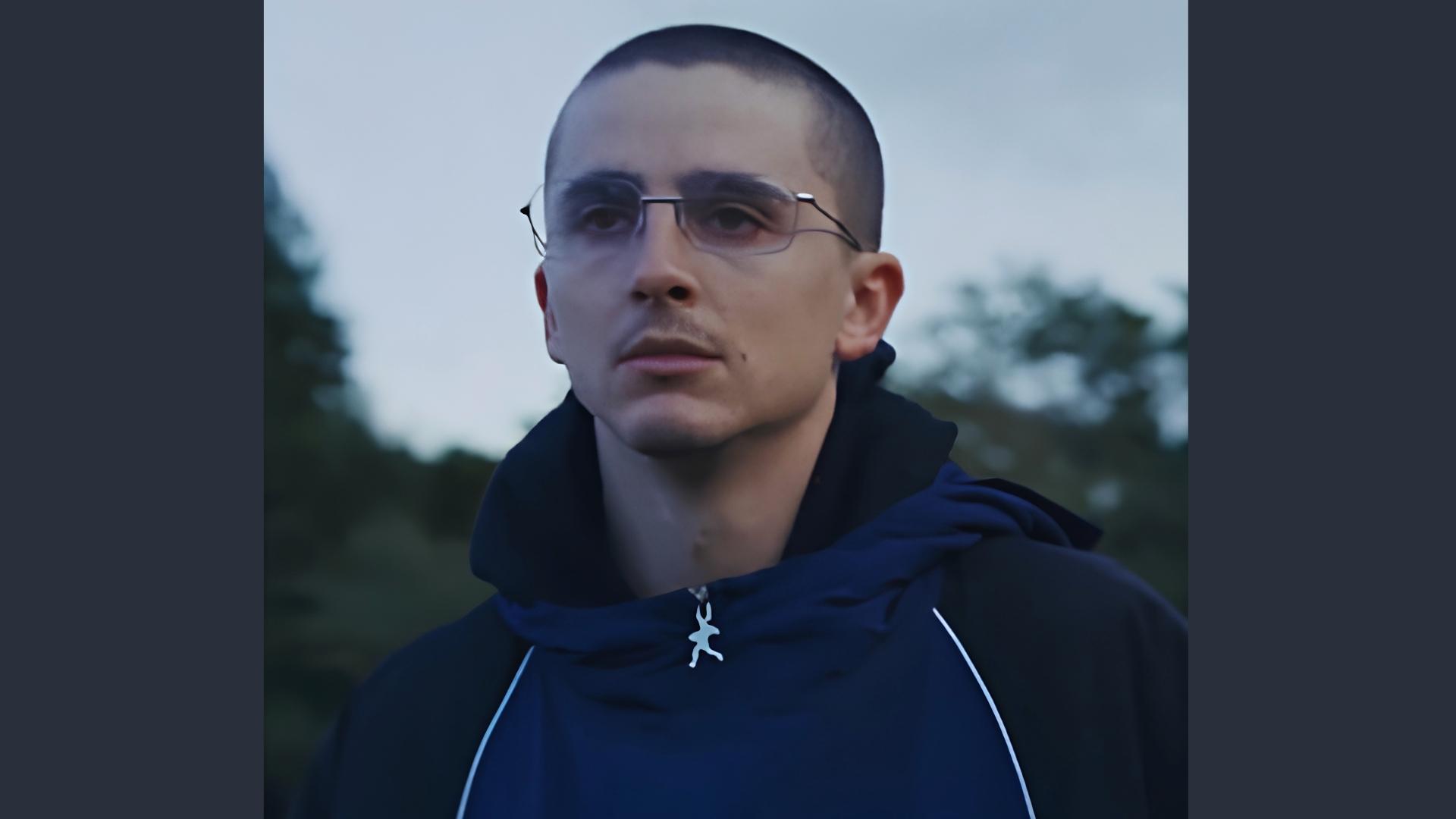 Timothée Chalamet with a tight military-style buzz cut and wire-rim glasses outdoors, wearing a dark hoodie with serious expression.
