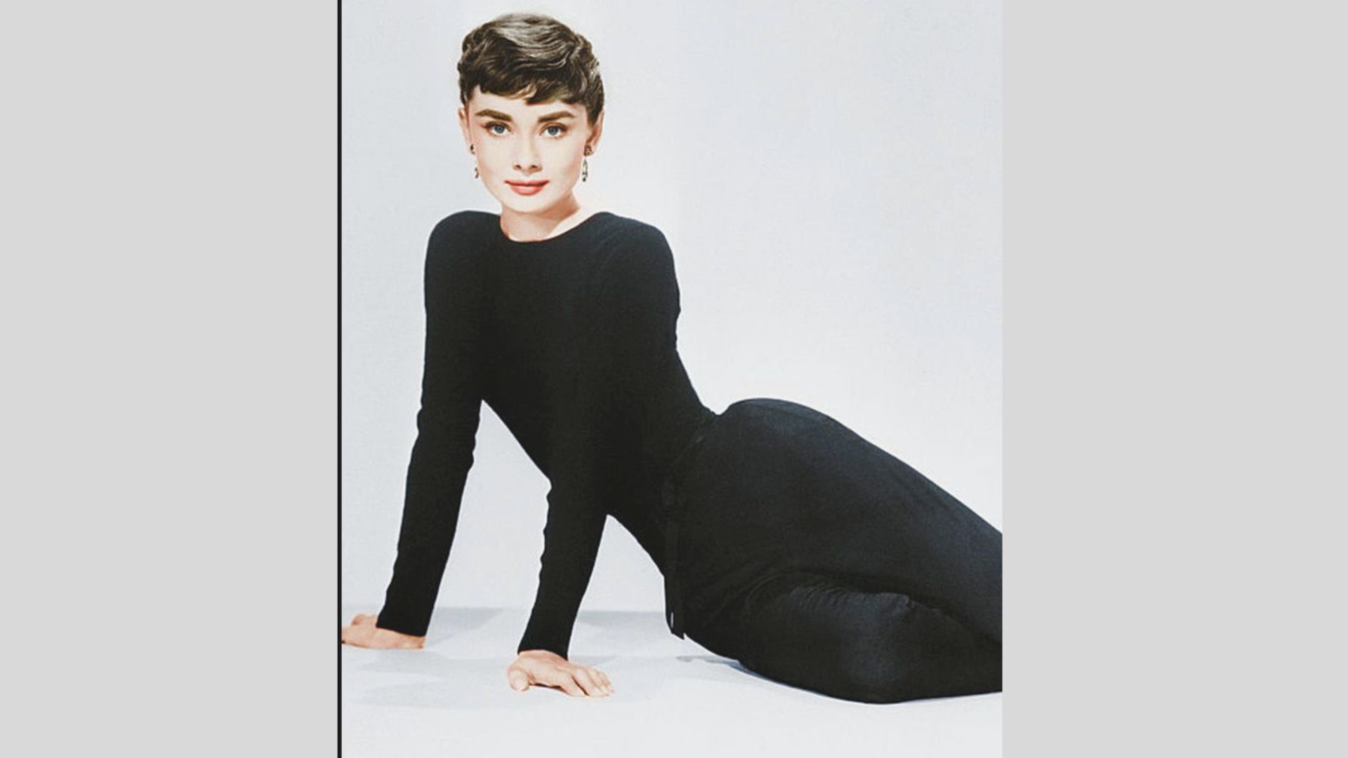 Color studio portrait of Audrey Hepburn in a fitted black long-sleeve boatneck top and slim black pants.