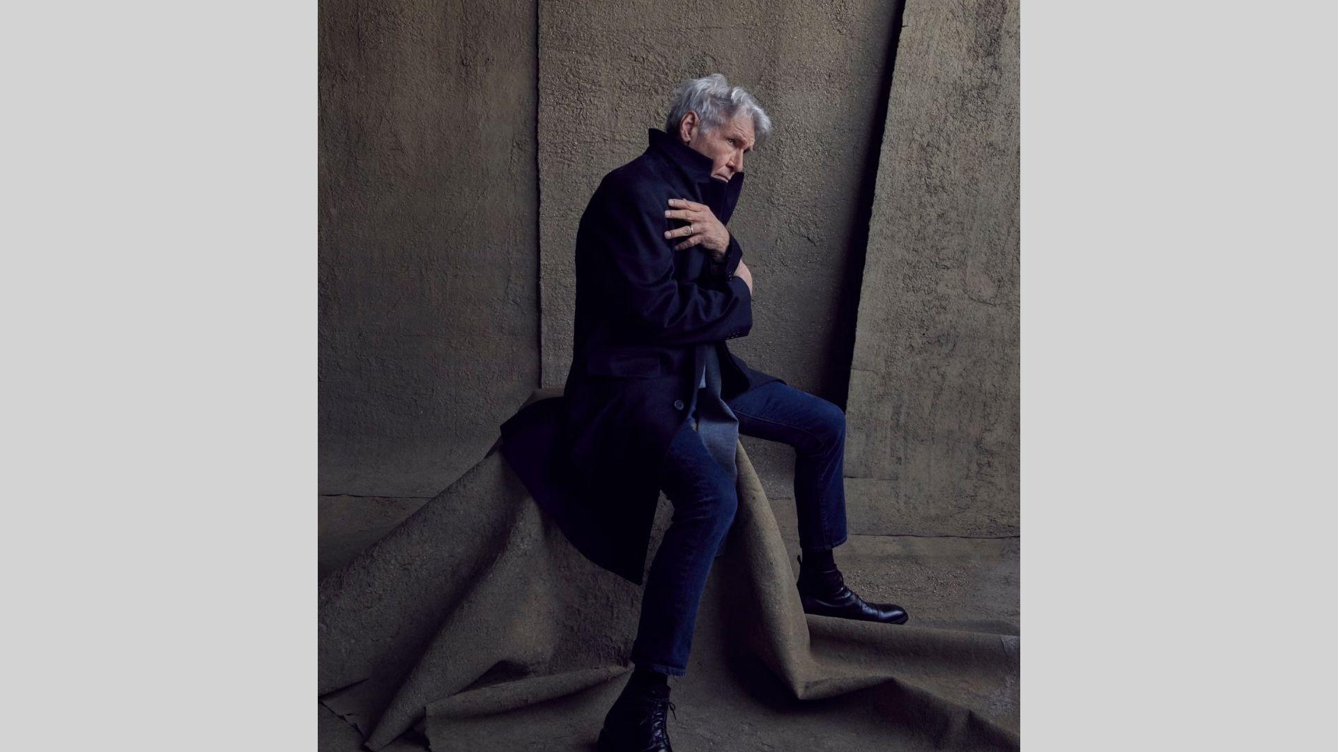 Older Harrison Ford sits hunched in black coat and jeans against textured concrete wall