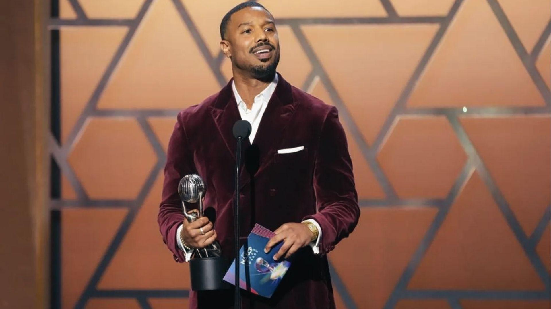 Michael B. Jordan holding award in burgundy velvet blazer over white shirt on stage.