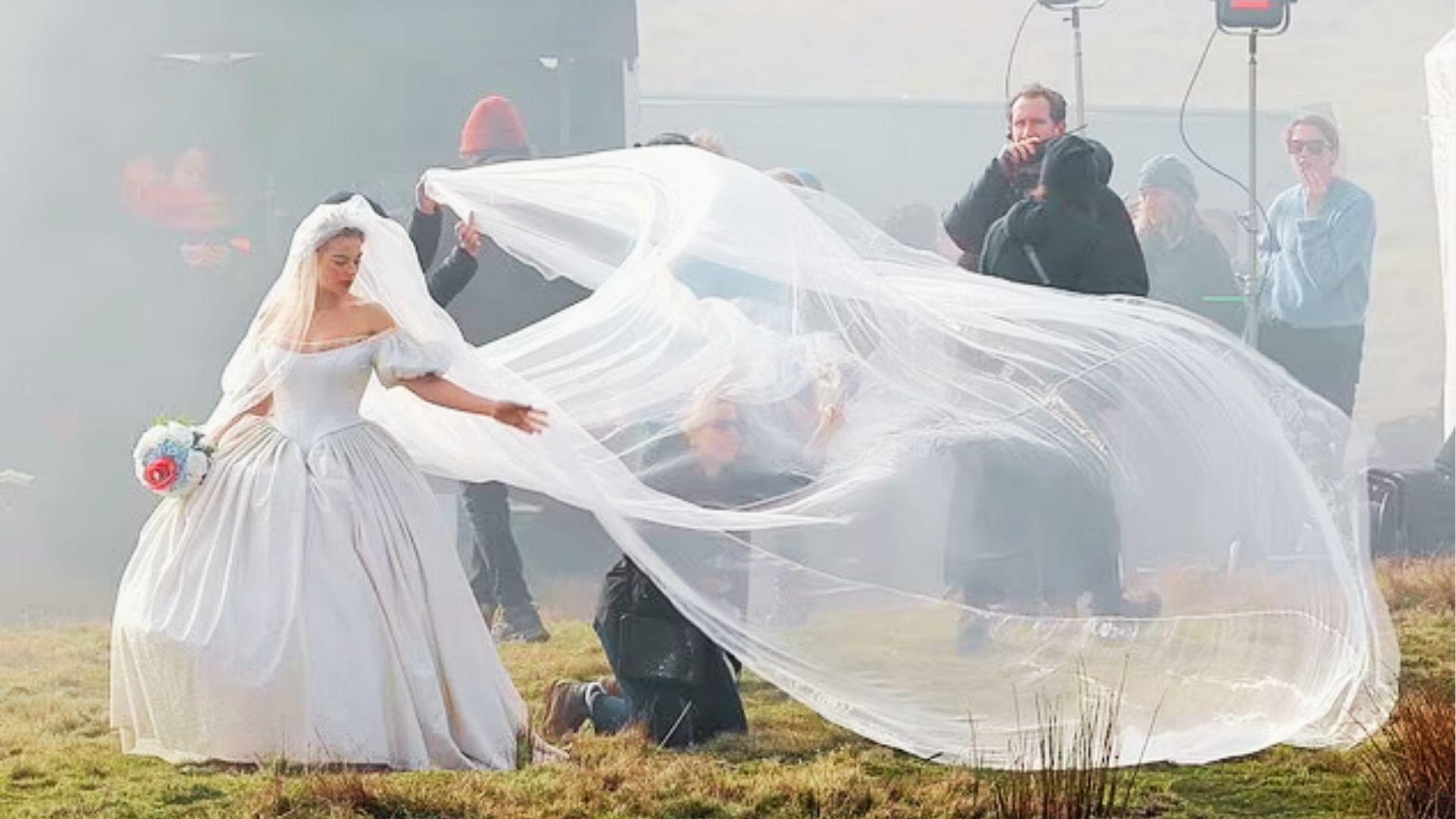 Bride in off shoulder gown holds bouquet while crew lifts flowing veil on windy film set
