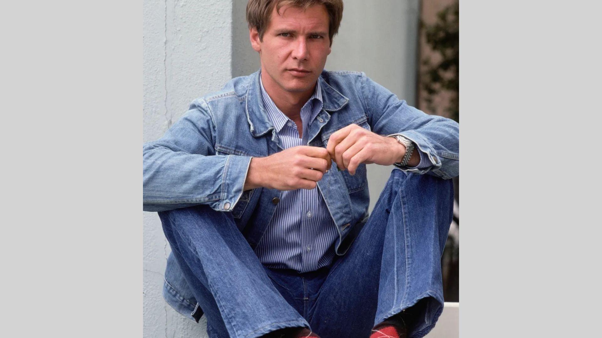 Young Harrison Ford sits cross-legged in denim jacket and jeans holding object while posing
