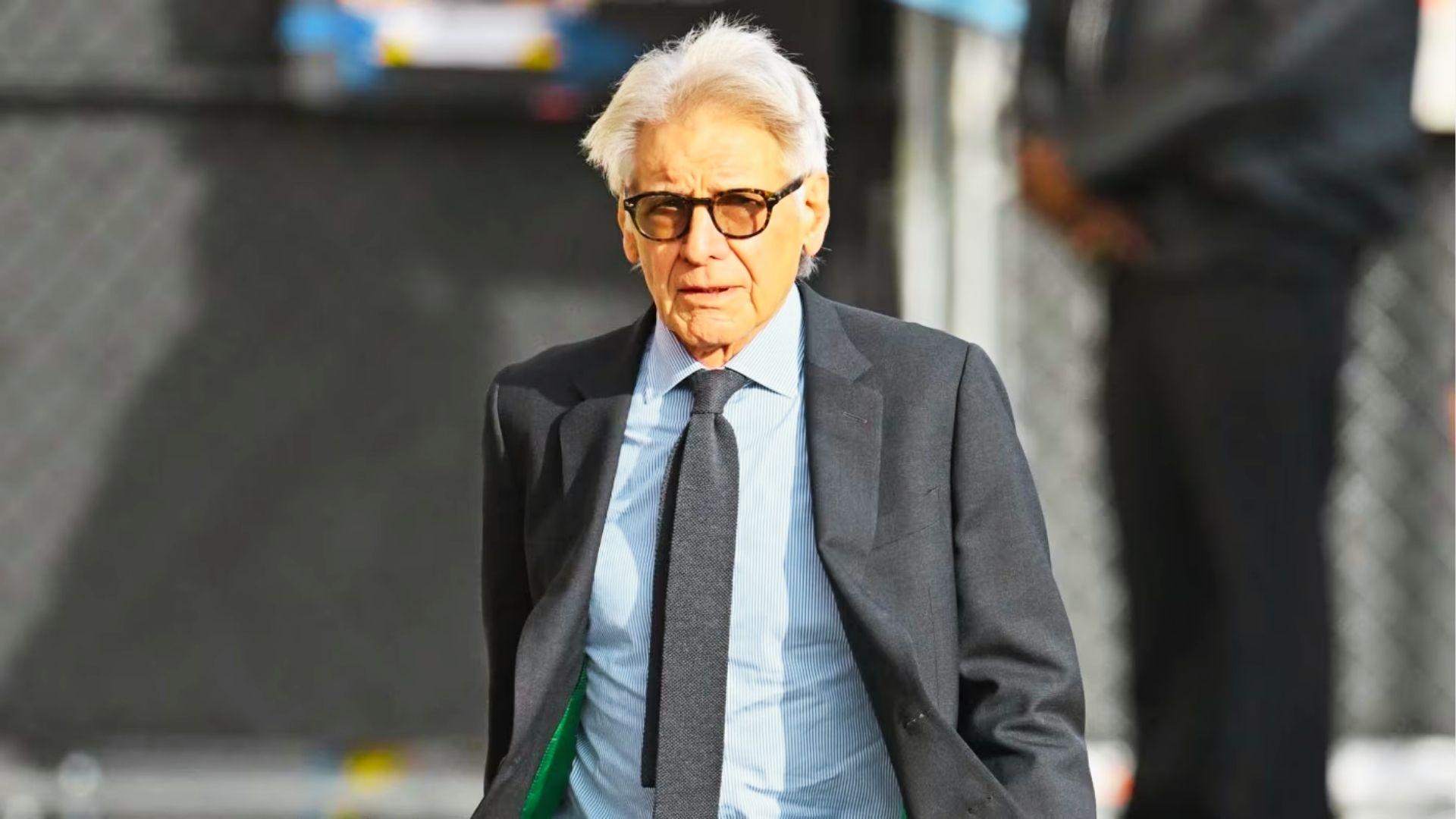 Older Harrison Ford walks outdoors in gray suit, blue shirt, dark tie, and glasses